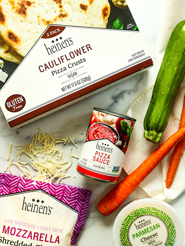 Spread of ingredients including Heinen's cauliflower pizza crusts, pizza sauce, shredded mozzarella cheese, zucchini, carrots, and parmesan cheese.