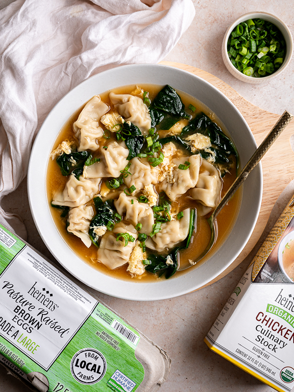 Egg drop soup in a bowl next to chives, a container of eggs, and box of chicken stock.