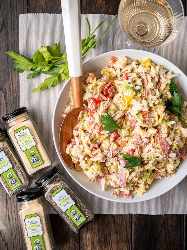 Grinder salad in a serving bowl with a wooden spoon, placed next to containers of Heinen's spices.