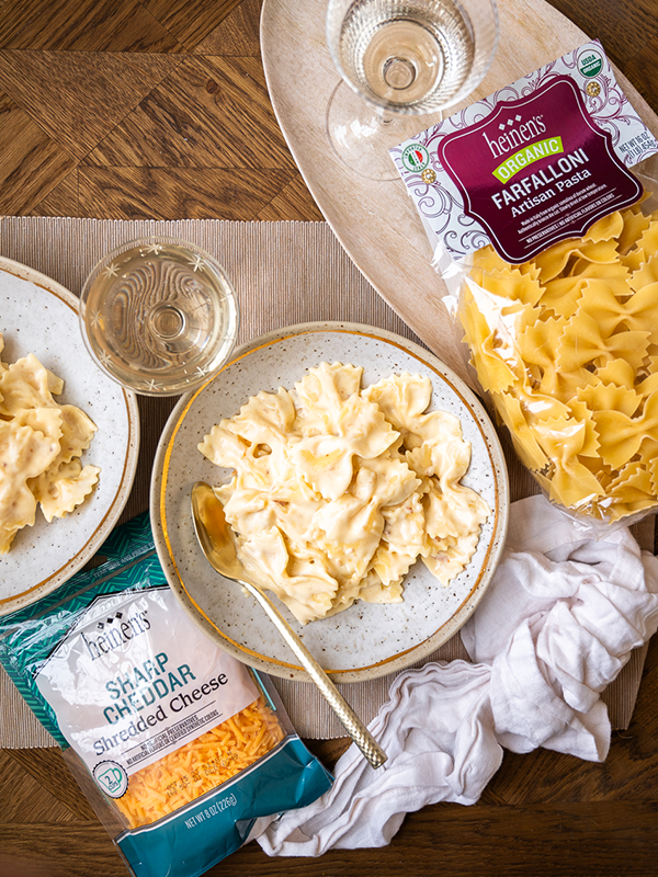 Farfalloni mac and cheese served in a bowl next to shredded sharp cheddar cheese, Farfalloni artisan pasta, and a glass of wine.