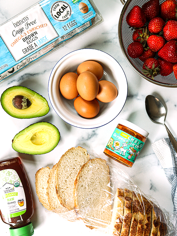 Kitchen counter with honey, sliced bread, avocados, eggs, strawberries, and chili oil.