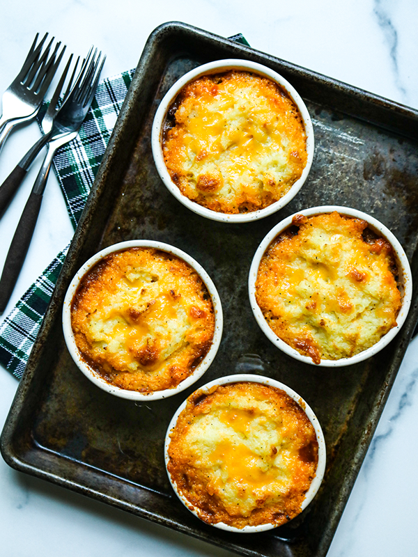 Individual cottage pies in ramekins on a baking sheet, topped with browned mashed potatoes.