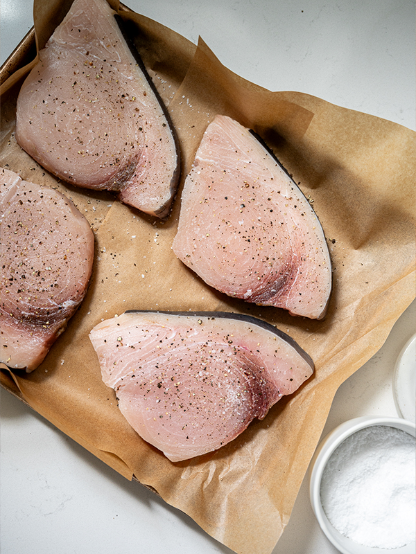 Raw swordfish on a baking sheet seasoned with salt and pepper.