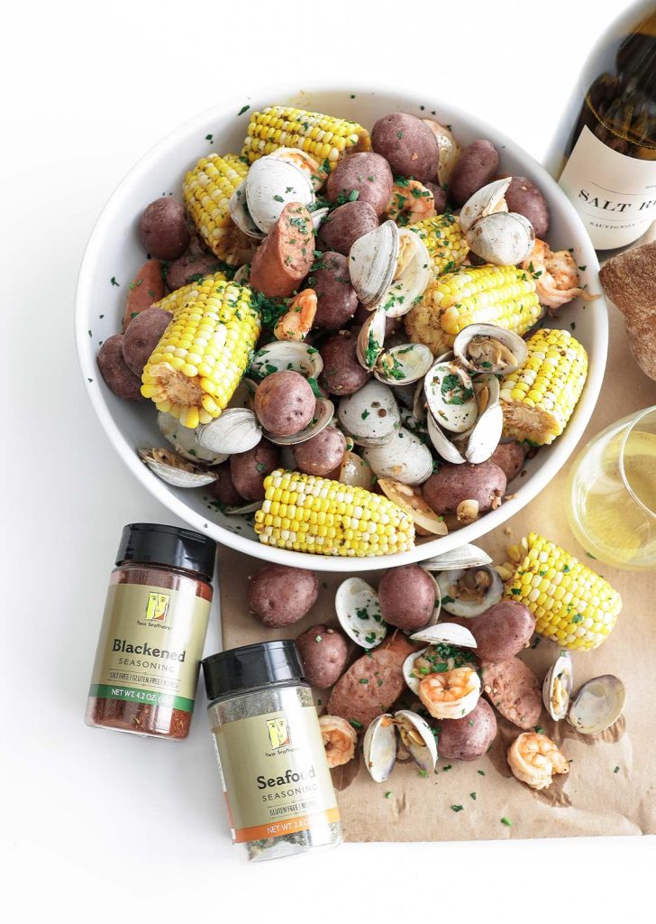 Large serving bowl filled with corn, clams, potatoes, sausage and shrimp next to seasonings and white wine.