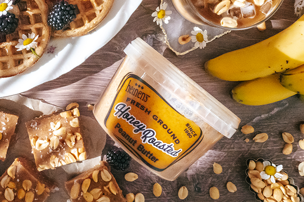 Container of Heinen's honey roasted peanut butter next to peanut butter squares, bananas, waffles, and an iced coffee.
