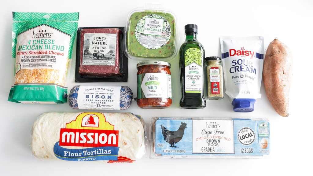 Table full of ingredients for Bison Breakfast Burritos including sweet potatoes, olive oil, Force of Nature ground bison, adobo seasoning, eggs, burrito shells, guacamole, sour cream, salsa, cheese, and cilantro.