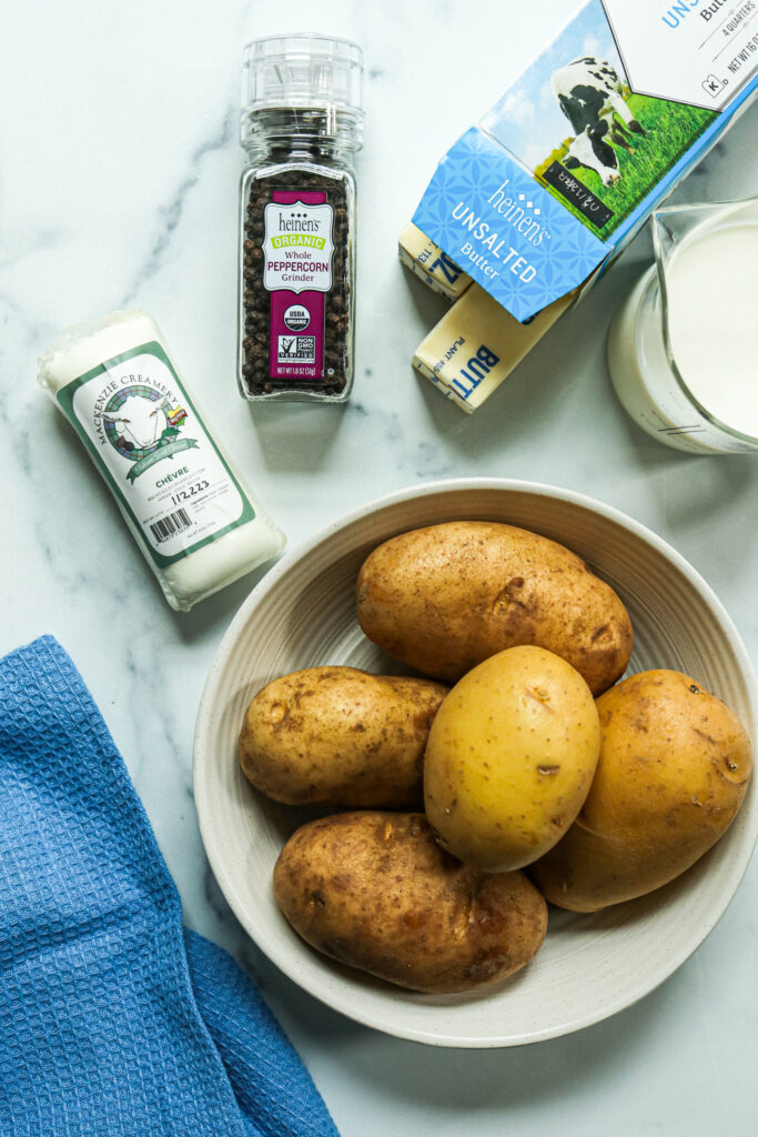 Freshly washed potatoes in a bowl next to milk, pepper, and unsalted butter.