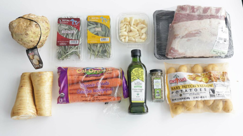 Pork roast ingredients arranged on a counter, including rosemary, sage, parsley, cloves, olive oil, carrots, parsnips, celery root, potatoes, and seasonings.
