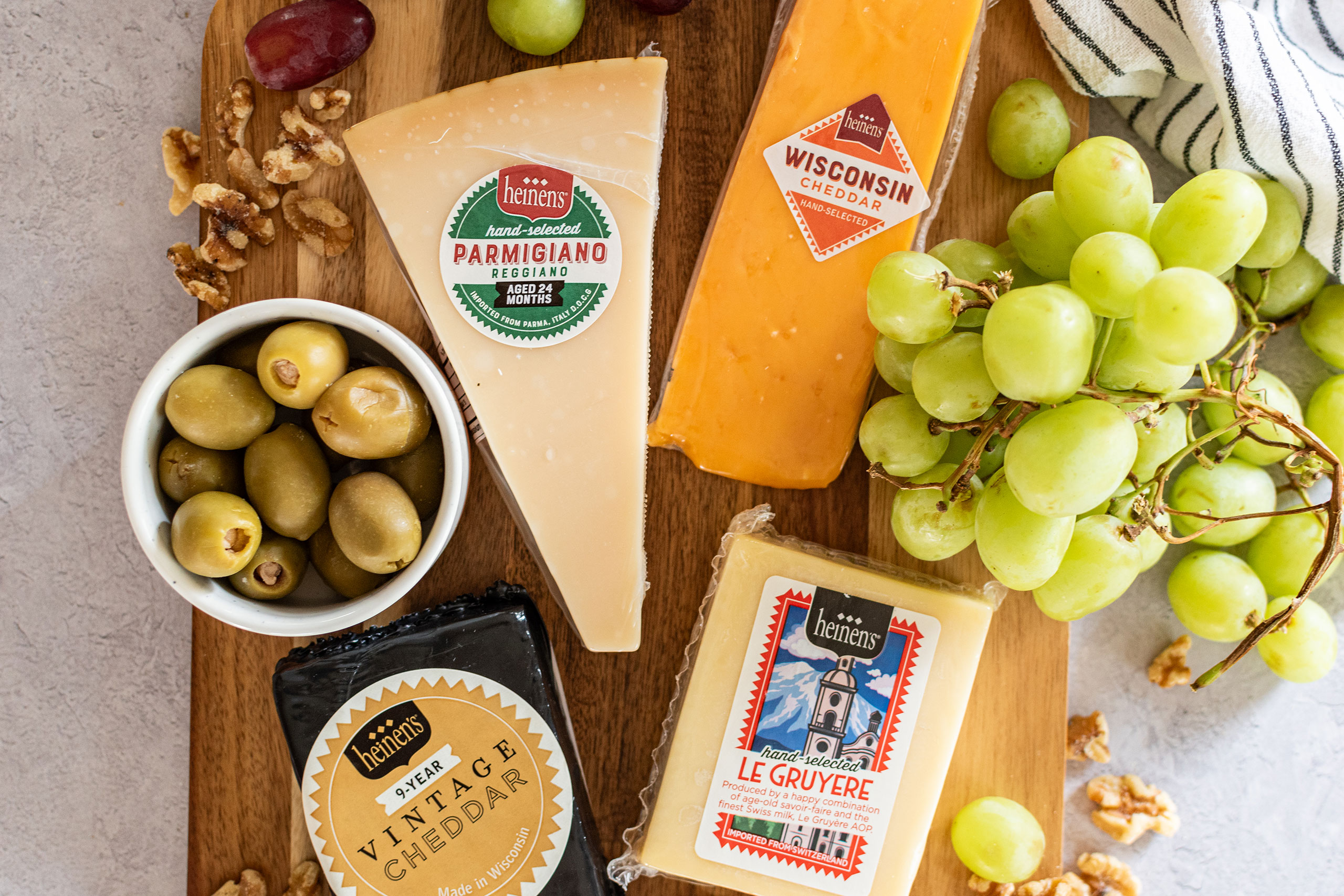 Heinen's Brand Hand-Selected Parmigiano, Wisconsin Cheddar, Vintage Cheddar, and Le Gruyere cheeses on a platter with green olives and green grapes.