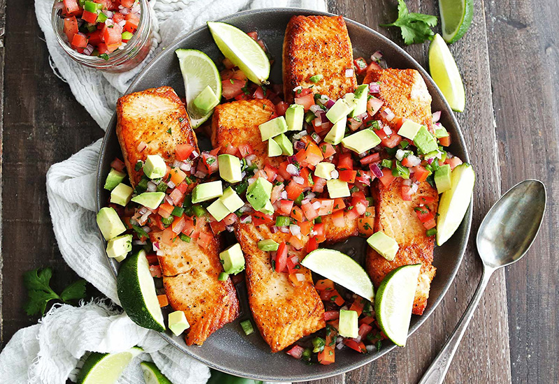 A plate of cooked salmon fillets topped with tomato salsa and chopped avocados, served with sliced limes. 