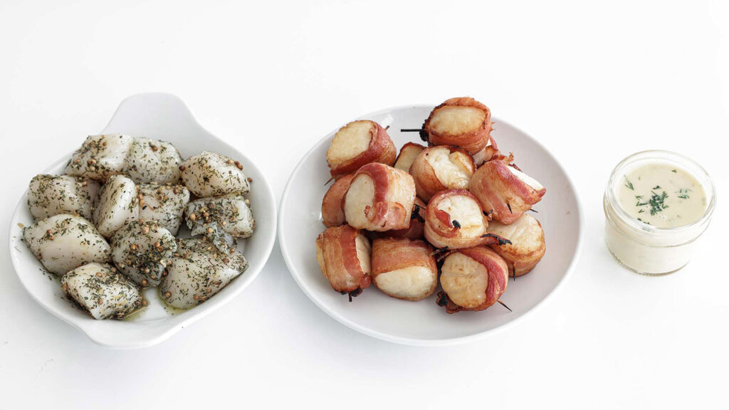 Bowls of scallops (seasoned and bacon-wrapped) arranged on a counter with a side of caper butter sauce.