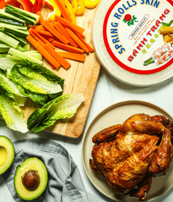 Roasted chicken on a plate next to lettuce, carrots, cucumbers, avocado, and spring roll wrappers.