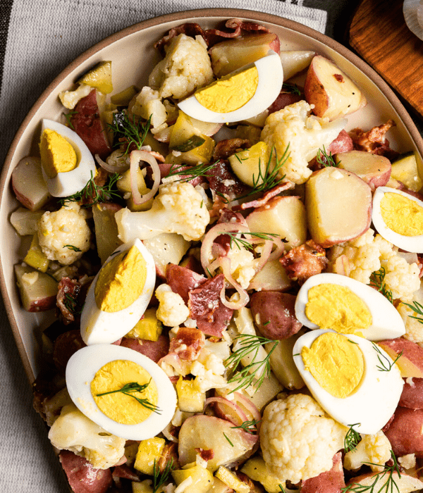 Close-up of creamy deli pickle potato salad with diced hard-boiled eggs, red potatoes, cauliflower, red onions, bacon, and a sprinkle of herbs.