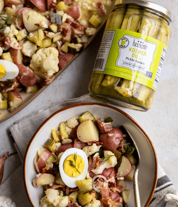 A bowl of deli pickle potato salad with a jar of Heinen's Kosher Dill pickle spears on its side next to the bowl.