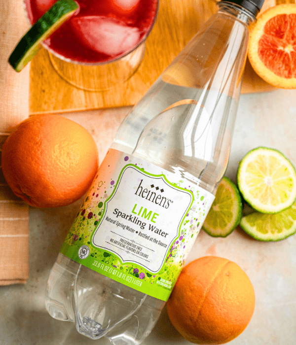 Bottle of Heinen's Lime Sparkling Water laying next to oranges & slices of limes.