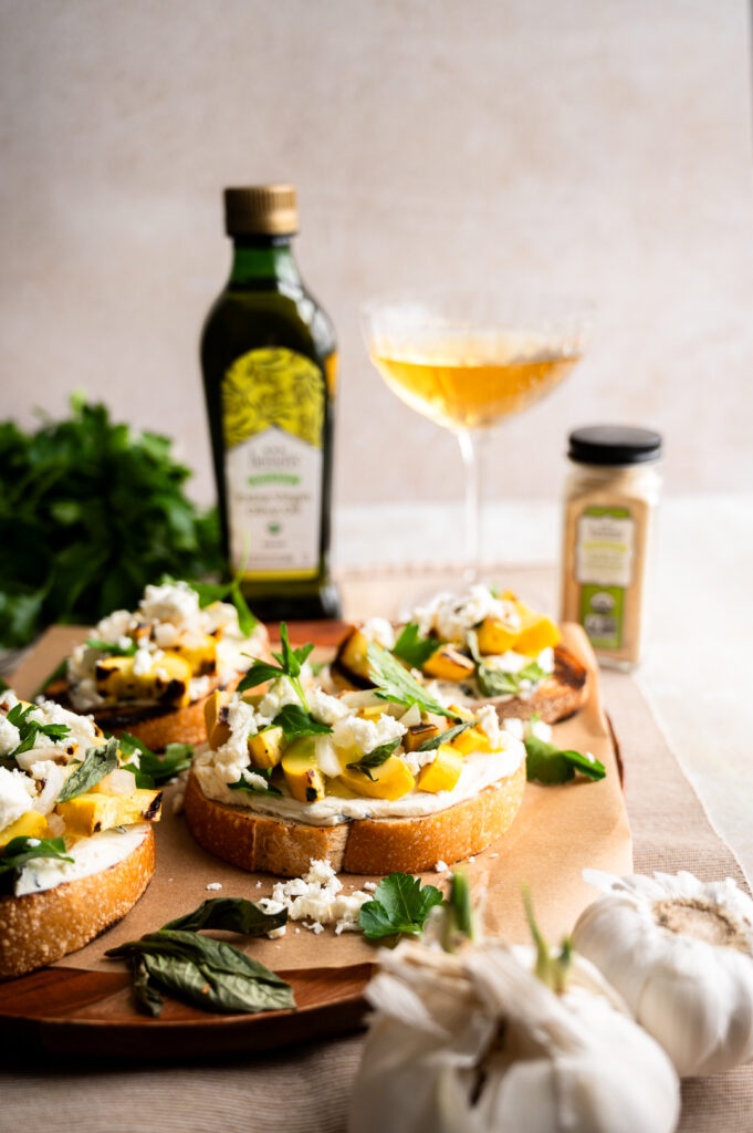Toast topped with feta cheese and yellow squash on a serving tray next to olive oil, fresh garlic, and seasoning container.
