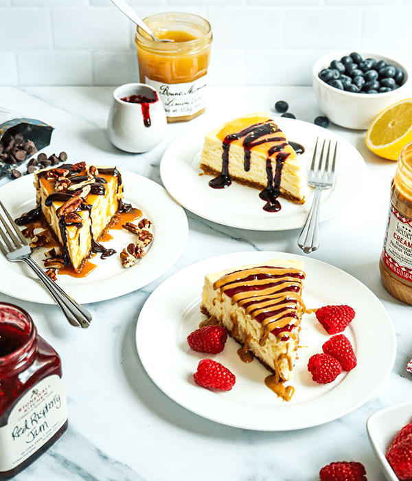 Vertical Image of Three White Plates Featuring Individual Slices of Heinen's Original Cheesecake with PB&J, Turtle and Lemon Blueberry Toppings. Individual containers if Topping Ingredients Sit in the Background.