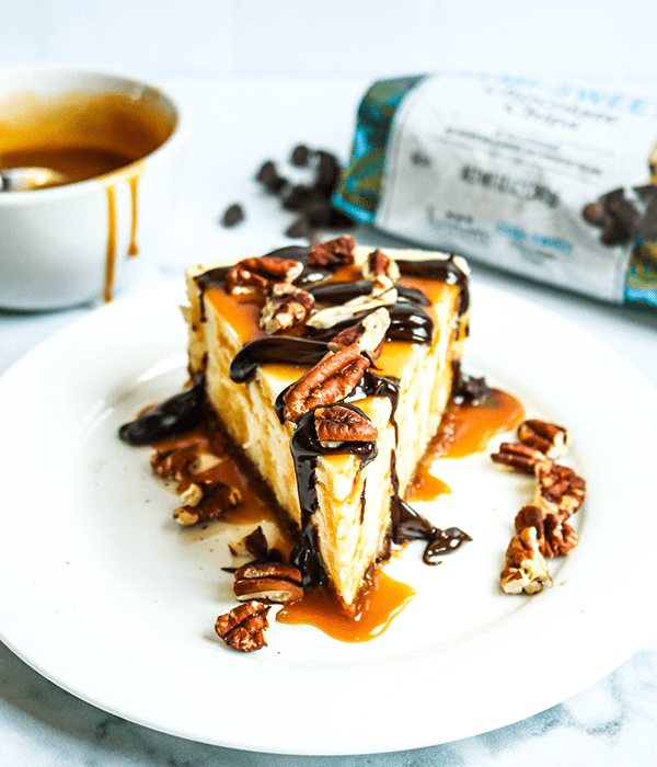 A Slice of Heinen's Original Cheesecake on a Plate with a Caramel, Chocolate and Pecan Turtle Topping. A Jar of Lemon Curd, a Bowl of Caramel Sauce and an Open Bag of Chocolate Chips Sit in the Background.