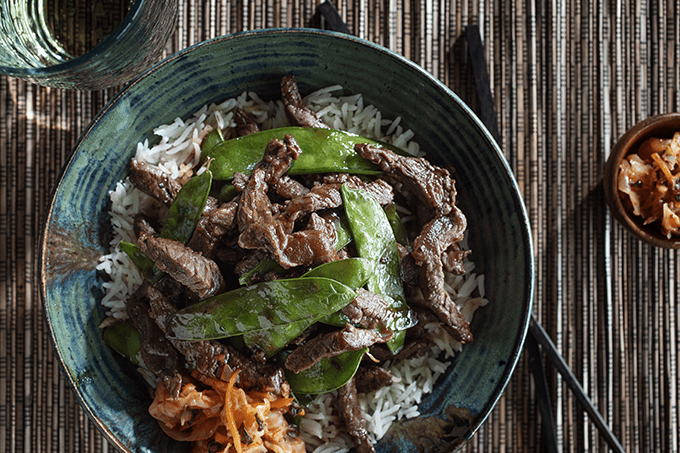 A bowl of rice topped with beef bulgogi, snow peas, and a spoonful of kimchi.