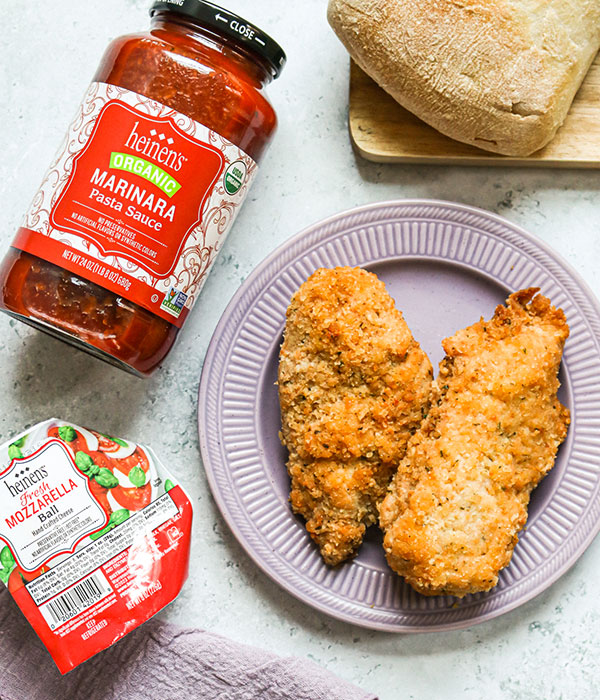 Two Pieces of Heinen's Chicken Romano on a Purple Plate Surrounded by a Jar of Heinen's Marinara Sauce and Heinen's Mozzarella Ball.