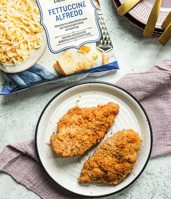 A Plate with Two Pieces of Heinen's Chicken Romano Beside a Bag of Heinen's Frozen Fettuccine Alfredo