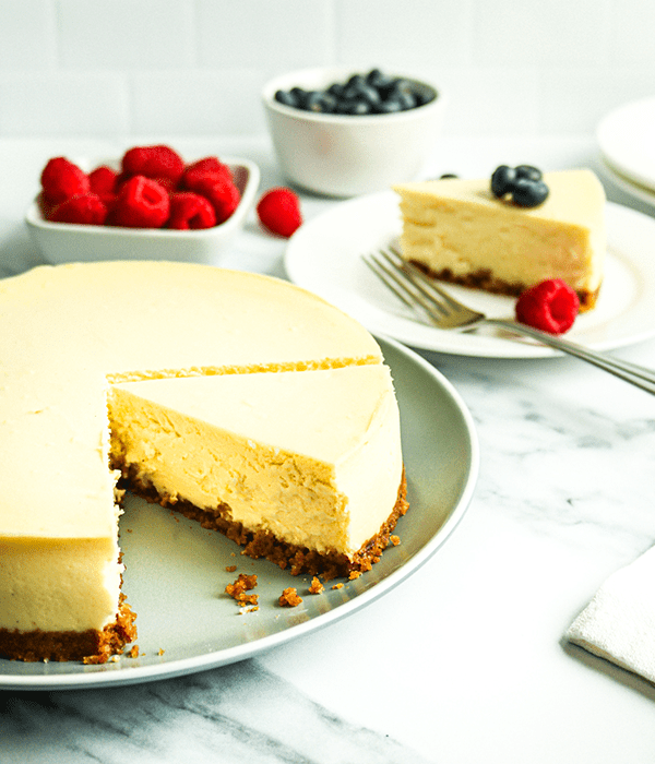 A Plain, Whole Heinen's Original Cheesecake on a Plate with a Slice Missing. A Plate with Single Cheesecake Slits sits in the Background with Bowls of Fresh Raspberries and Blueberries.