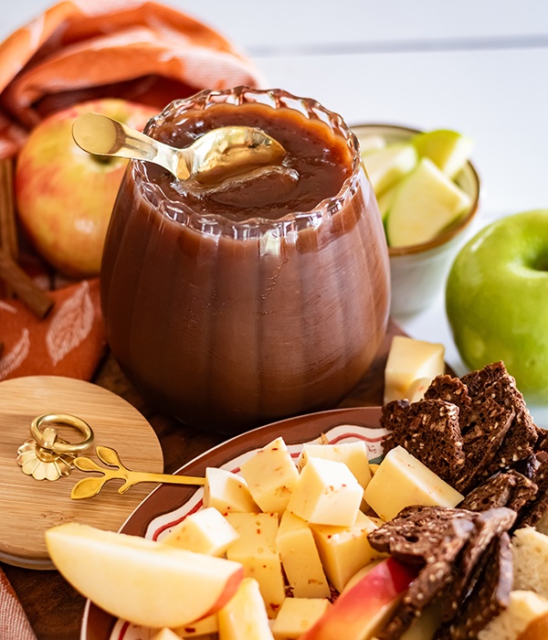 A jar of homemade apple butter with a gold spoon, surrounded by apple slices, cheese cubes, and crackers on a serving board.