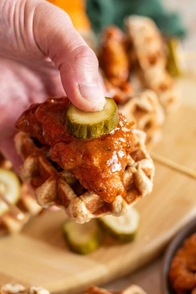 A hand holding a mini waffle topped with Nashville hot chicken and a bread and butter pickle.