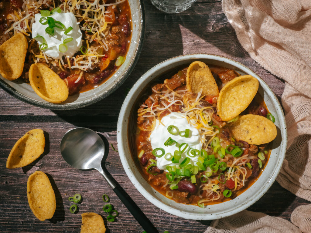 Two bowls of red chili topped with sour cream, cheese, green onions and Fritos.