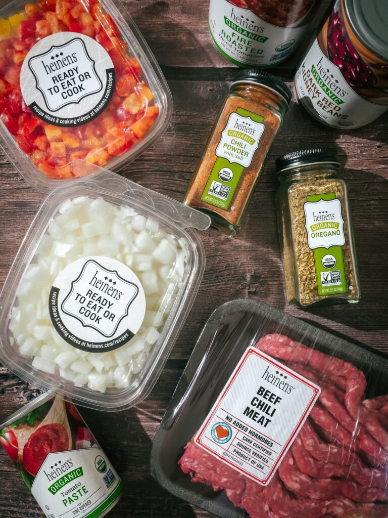 An overhead view of Classic Beef Chili ingredients, including containers of chopped onions, bell peppers, tomato paste, ground beef, various seasonings, and canned beans.