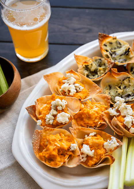 Daily Bites Spinach and Artichoke and Buffalo Chicken dips in baked wontons served on a plate with celery and blue cheese crumbles.