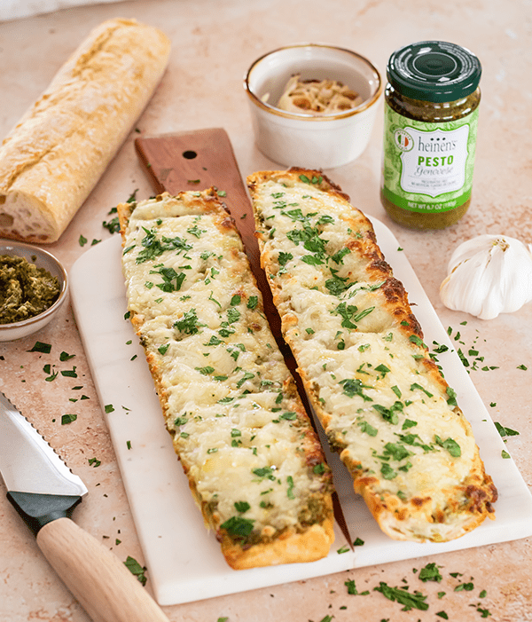Cheesy Pesto Garlic Bread loafs on a cutting board sprinkled with parsley and surrounded by a jar of pesto and a whole roasted garlic bulb.