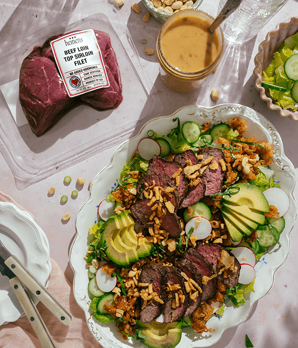 Romaine lettuce, cucumber, avocado and radishes topped with sliced steak and crispy rice on a serving plate, next to tahini salad dressing and Heinen's beef loin top sirloin fillet.