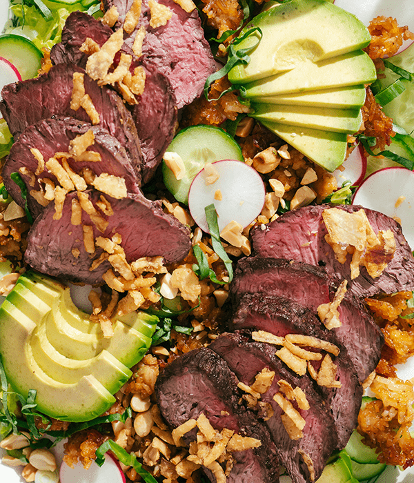 Romaine lettuce, cucumber, avocado and radishes topped with sliced steak and crispy rice on a serving plate,