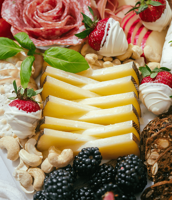 A Close Up Image of a Patriotic Charcuterie Board Featuring Triangle Cheese Slices, Red Berries, Blackberries, White Chocolate Covered Strawberries, Rolled Deli Meats, and Nuts