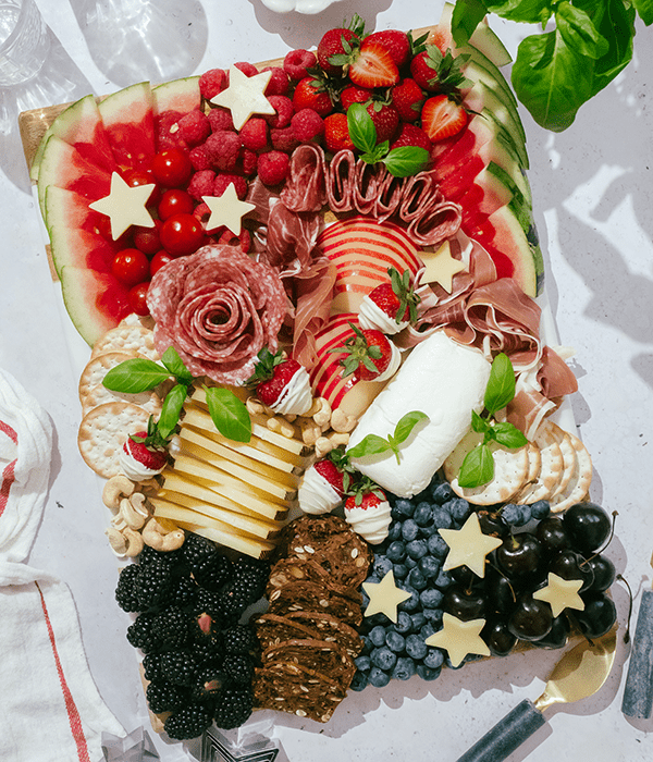 A Fully Assembled Charcuterie Board with Sliced Cheeses, A Whole Goat Cheese Log, Sliced Fruit, Rolled Charcuterie Meats, Red Berries, Blackberries, Blueberries, Cherries, Star-Shaped Cheese Slices, White Chocolate Covered Strawberries, and Fresh Basil.