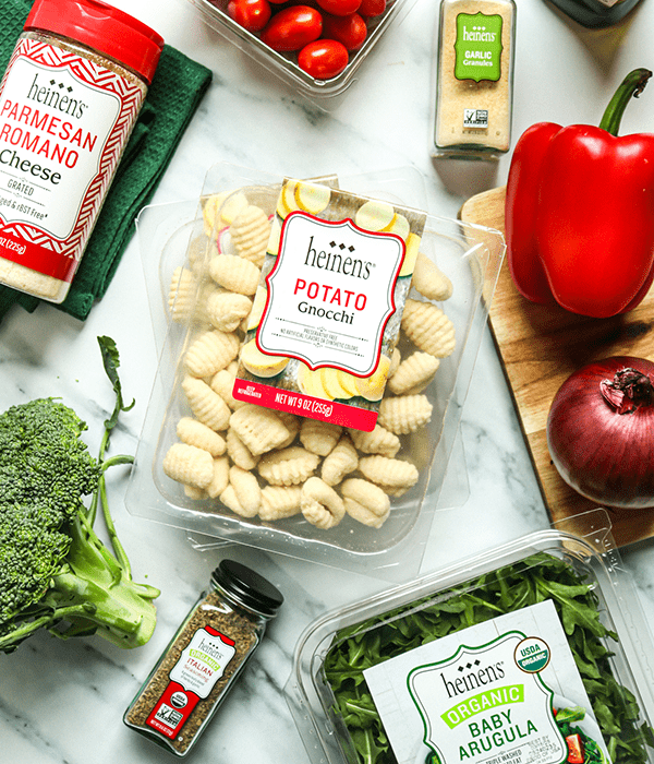 Package of Heinen's potato gnocchi next to garlic powder, parmesan romano cheese, broccoli, a red onion, a red bell pepper, and a package baby arugula. 