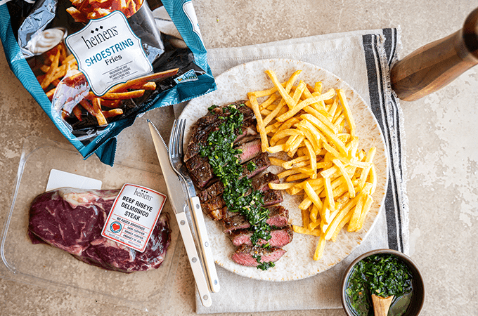 Sliced ribeye steak topped with chimichurri sauce and served with French fries, next to a packaged ribeye steak and bag of Heinen's shoestring French fries.