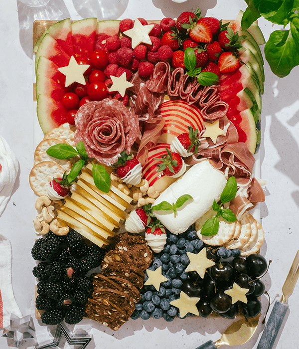 A Partially Assembled Charcuterie Board with Sliced Cheeses, A Whole Goat Cheese Log, Sliced Fruit, Rolled Charcuterie Meats, Red Berries, Blackberries, Blueberries, Cherries, Star-Shaped Cheese Slices, and White Chocolate Covered Strawberries.