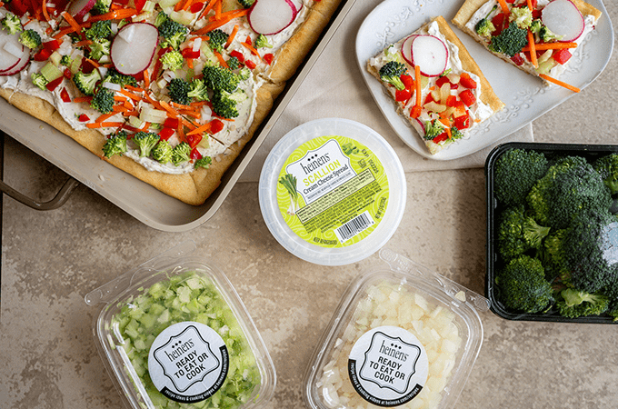 Cold veggie pizza on a baking sheet next to packages of Heinen's scallion cream cheese, pre-chopped celery and pre-chopped onion.