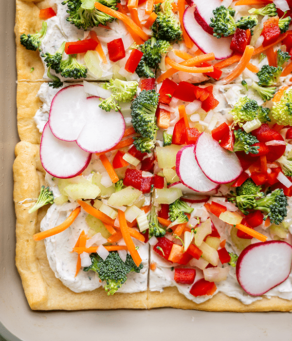 Square slices of pizza with cream cheese topped with radishes, broccoli, onion, bell pepper and cheese.