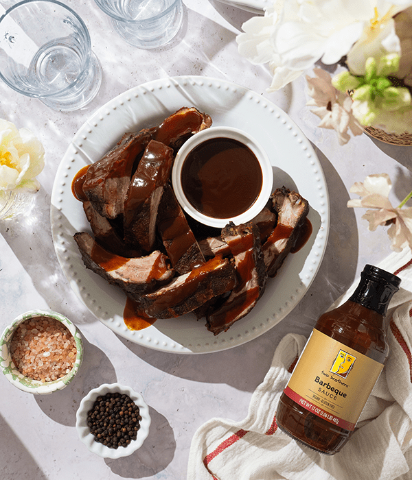Baby back ribs arranged on a serving plate, generously drizzled with BBQ sauce, displayed beside a container of Two Brothers BBQ Sauce.