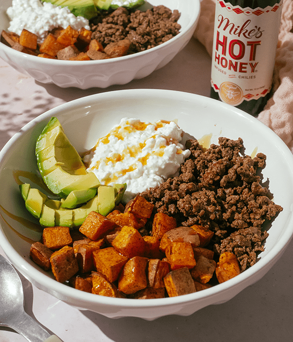 A Big White Serving Bowl with Ground Beef, Cottage Cheese, Sweet Potatoes, and Avocado Beside a Bottle of Mike's Hot Honey