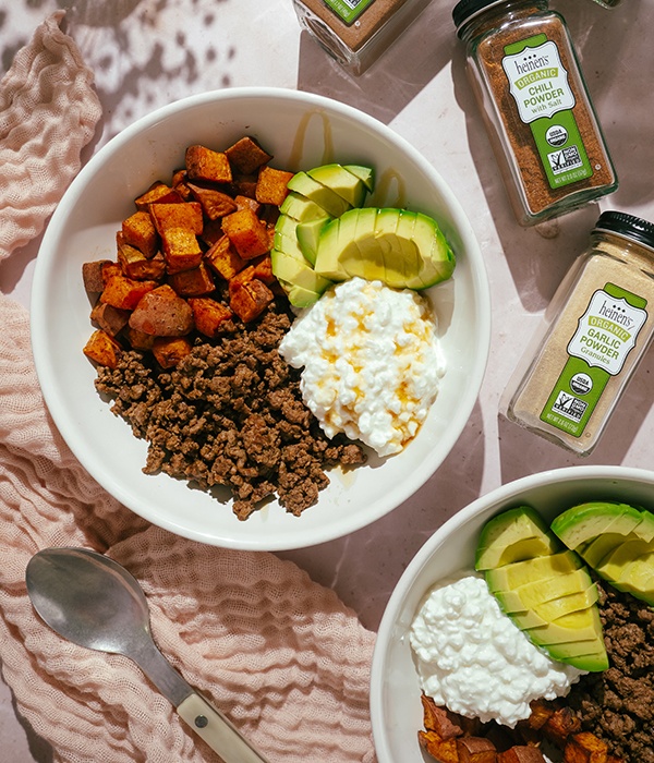 A White Serving Bowl Filled with Ground Beef, Sweet Potatoes, Avocado, and Cottage Cheese. Bowls are surrounded by spice jars.