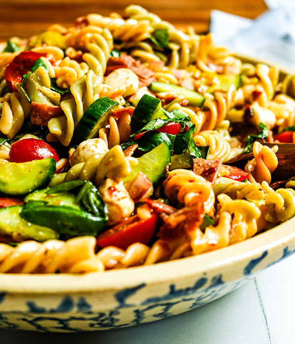Bowl of rotini pasta with tomatoes, cucumbers, onion, mozzarella, salami, pepperoncini, and prosciutto