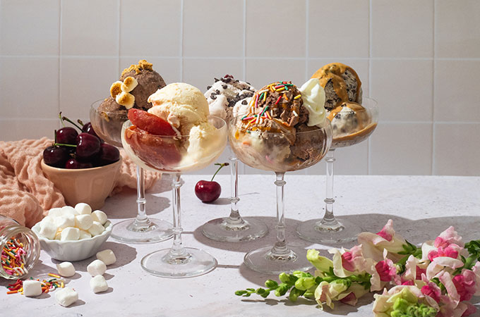 Five ice cream sundaes in dessert glasses, flowers, marshmallows, and cherries on side