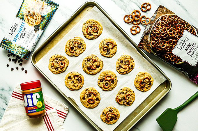 A cookie tray with parchment and peanut butter pretzel cookies. A bag of Heinen's pretzels and chocolate chips and a jar of Jif peanut butter.